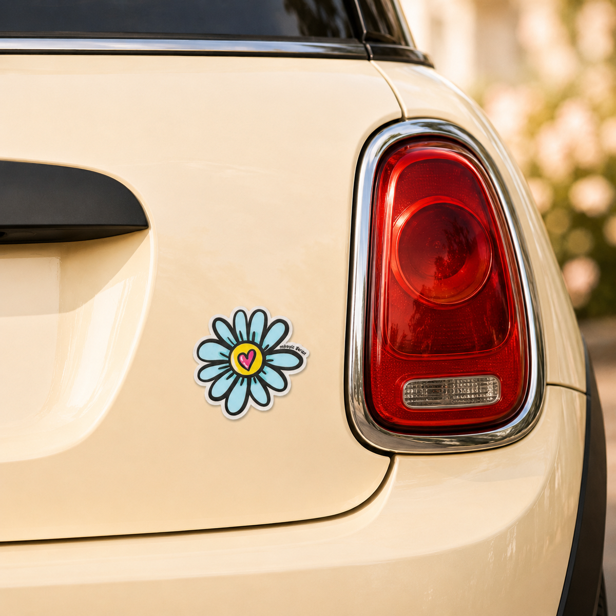 A die-cut flower sticker with a blue daisy-like outline, yellow center, and small pink heart is shown applied to the rear quarter panel of a cream-colored car next to the red taillight.